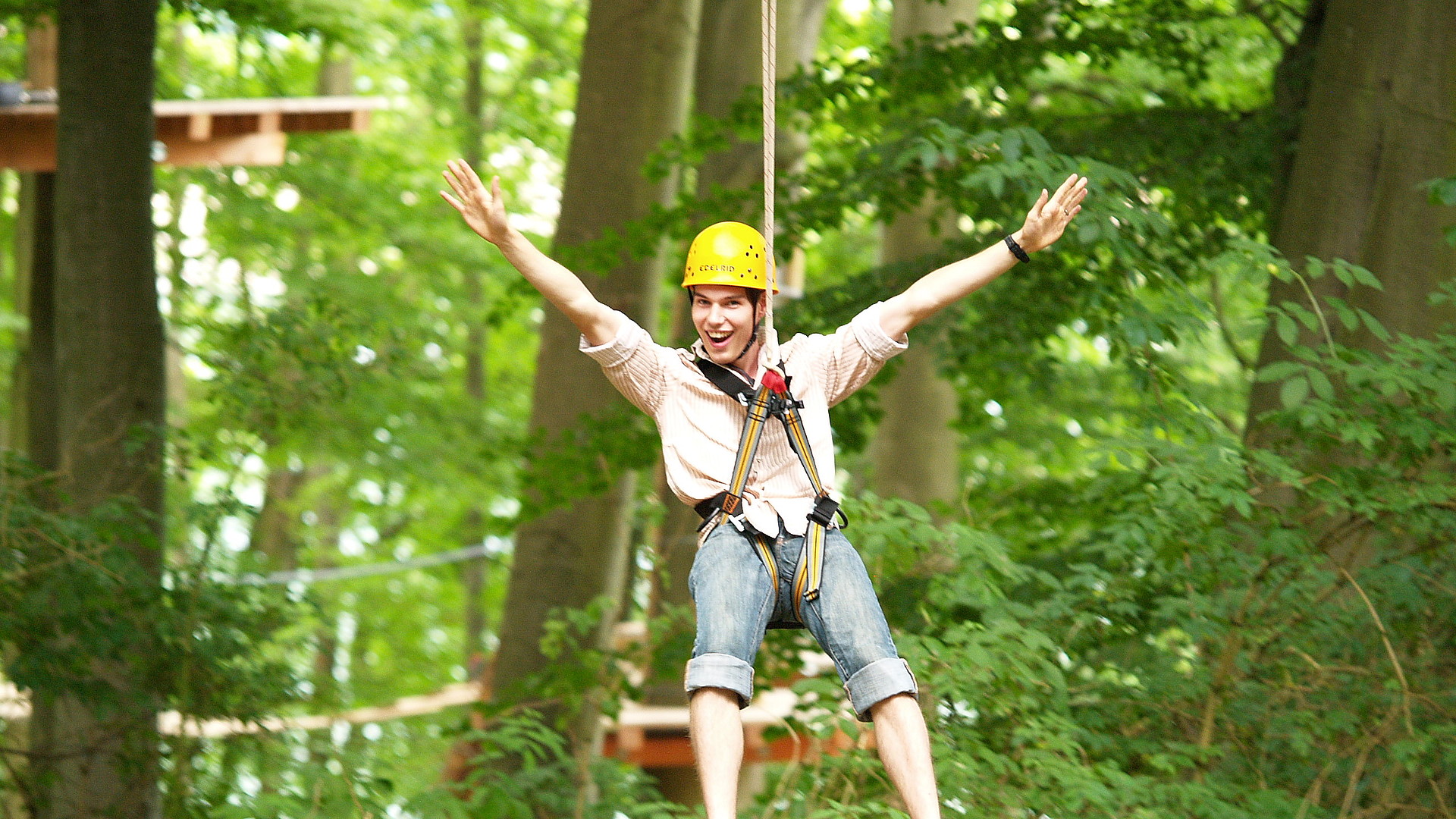 Junger Mann, der lacht und freudig die Arme in die Luft streckt, im Kletterwald auf einer Seilrutsche